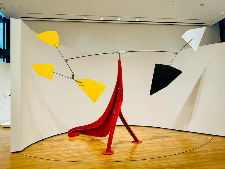 A large metal sculpture fills much of a room. Its base is a bright cherry red tripod shape. From the top sprouts a series of antennae with large yellow, black, and white paddles on the ends. There is something about the structure that references the circus.